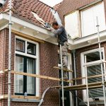 Maison bruxelloise en rénovation avec un artisan inspectant la toiture, les fenêtres et les équipements techniques à la lumière naturelle.